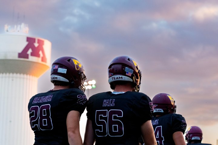 A group of football players wearing maroon helmets and black jerseys stand on the field, facing away from the camera. The sky above is painted with warm sunset colors. In the background, a water tower with the University of Minnesota 'M' logo is visible.